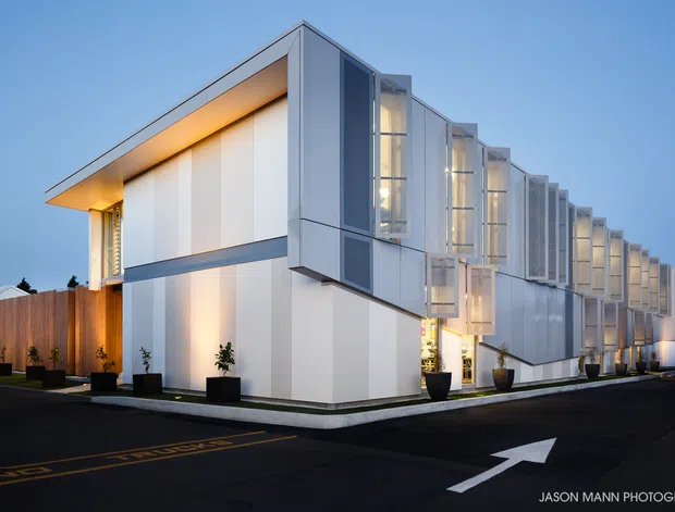 Exterior dusk photograph of a long, contemporary multi-storey building with a sharply folded roofline, white and grey panelled facade, extensive vertical glazing, projecting window bays, and warm architectural lighting. The building fronts a paved access road with directional markings and planted entry landscaping.