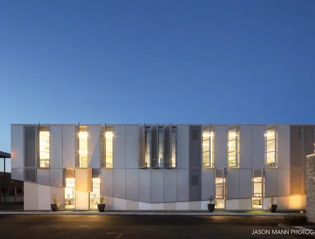 Twilight exterior view of a contemporary multi-storey building with a rectilinear form, pale panelled facade, extensive vertical glazing, recessed illuminated windows, and a sheltered entrance. The building fronts a paved area and is shown against a clear blue sky.