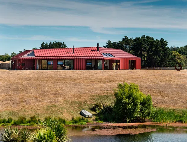 A single-storey red corrugated-metal-clad building with a low pitched roof sits in an open rural landscape. Large glazed openings line the facade, with several roof skylights or glazed roof sections and visible flues. A pond and dense native-style planting occupy the foreground, while dry grassland and mature trees surround the building under a broad sky.