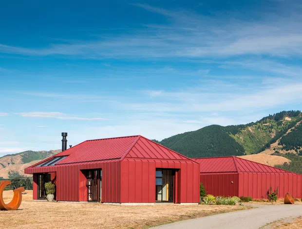 Exterior view of a low, single-storey red metal-clad building with multiple pitched standing-seam roof forms, large glazed openings, and a connected lower wing. The building sits in a dry landscaped setting with rugged, forested hills and mountains behind it. A sculptural orange outdoor object and sparse planting appear near the entrance.