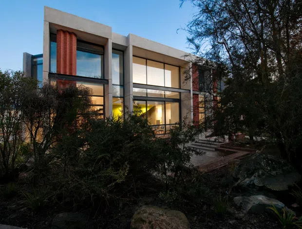 Exterior view of a contemporary two-storey residence set within dense landscaped gardens. The building features a rectilinear concrete frame, extensive floor-to-ceiling glazing, dark window frames, warm interior lighting, and prominent red textured vertical wall elements. Stone-edged landscaping and steps lead toward the house.