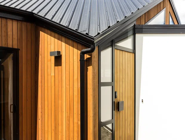 Exterior architectural photograph of a compact contemporary building with warm vertical timber cladding, dark metal-framed glazing and a standing-seam metal gable roof. A black downpipe and gutter are prominent, with a timber deck and pale paved ground in the foreground under a bright blue sky.