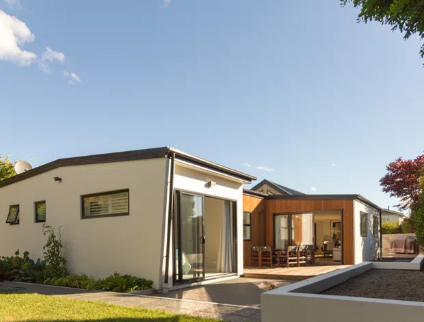 Exterior view of a contemporary single-storey residential extension or compact home, with white rendered walls, dark trim, broad glazed sliding doors, and a connected timber-clad pavilion. The building sits in a landscaped suburban garden with paved outdoor areas and a tall timber boundary fence, photographed in bright daylight.