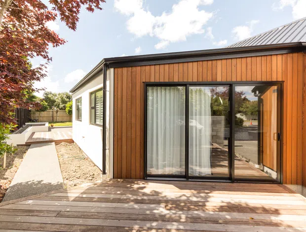 Exterior view of a modern single-storey home or extension, featuring warm vertical timber cladding, black-framed sliding glass doors, a low-pitched metal roof, and a timber deck leading to a paved side path and courtyard-like outdoor area. The interior is only partially visible through the glazing.