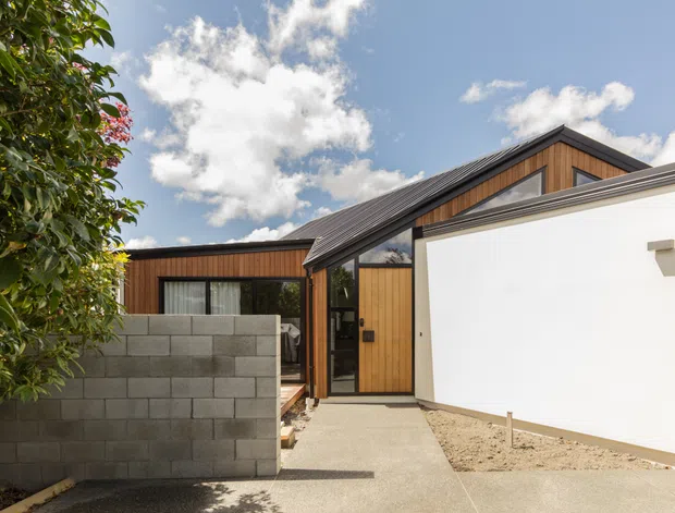 Exterior view of a contemporary low-rise residence with a gabled roof, dark metal roofing, warm vertical timber cladding, white rendered walls, and a concrete block boundary wall. A concrete walkway leads to the recessed timber entry door, with landscaped planting at the edge of the frame under a bright blue sky.