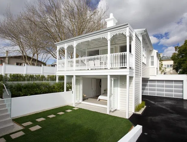 Exterior view of a refined two-storey white weatherboard home with an ornate covered veranda and balcony, decorative fretwork, balustrades, columns, and a small chimney. The lower level opens toward a compact landscaped lawn, while a dark paved driveway leads to an attached double garage. Neighboring homes, fencing, stairs, and mature trees are visible around the property.