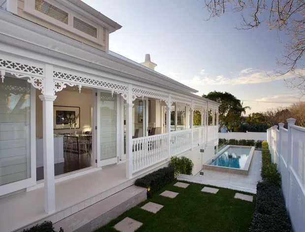 Exterior view of an ornate, light-painted residence with a long covered veranda featuring decorative fretwork, turned-style posts, balustrades, and glazed doors. A manicured courtyard garden with stepping stones and a narrow rectangular pool runs alongside the house, enclosed by a white privacy fence. The image has a refined heritage-inspired character with bright, warm evening light.