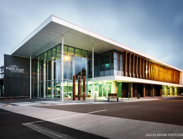 Exterior dusk photograph of the Kapiti Coast District Council building, featuring a contemporary two-storey civic structure with a dramatic cantilevered flat roof, extensive floor-to-ceiling glass, dark solid wall cladding, and a prominent timber-toned vertical sun-screen facade. The building fronts a paved pedestrian plaza and driveway.