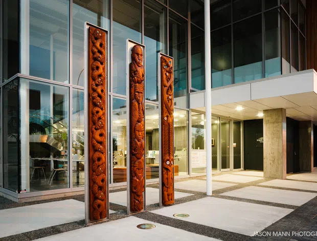Contemporary multi-storey building entrance with extensive floor-to-ceiling glazing, a projecting white canopy, exposed concrete columns, and dark cladding. Three tall carved timber sculptural panels stand prominently in the forecourt, with patterned light paving and dark gravel bands leading toward the glazed lobby.