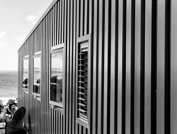Black-and-white exterior architectural photograph of a long, narrow building clad in vertically ribbed dark metal panels. A series of framed windows and a louvred window punctuate the facade, which is elevated on slender supports above a sloping coastal site with the ocean visible beyond.