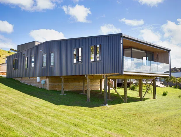 Exterior view of a contemporary elevated single-storey home on a sloping grassy site. The building features dark vertical metal cladding, a low-pitched roof, timber substructure and screening, narrow vertical windows, and a large glazed balcony or outdoor living area overlooking the water. Bright daylight and expansive glazing create a modern coastal setting.