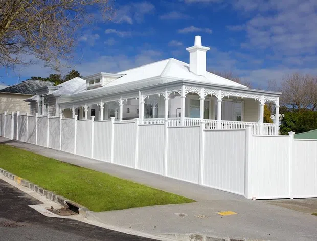 Street-side view of a renovated or newly built single-storey white house with a hipped metal roof, prominent chimney, enclosed front verandah, ornate decorative posts and fretwork, white balustrades, and a tall white picket-style boundary fence. The home sits on a gently sloping suburban site with a driveway and lawn.