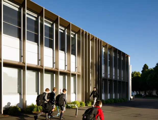 Exterior view of a contemporary multi-storey institutional or commercial building with a long rectilinear facade, repeated full-height glazing, white wall panels, and prominent vertical dark metal fins. Students and a paved forecourt appear in front, with trees at the edge of the site under a clear blue sky.