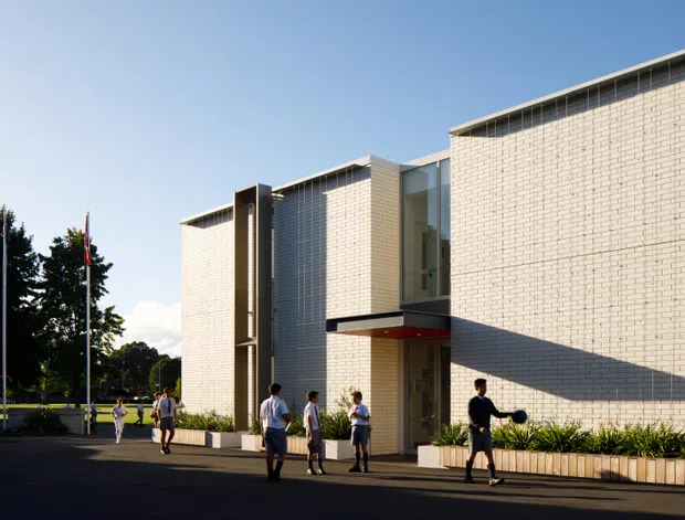 Exterior architectural photograph of a contemporary institutional building with pale textured masonry walls, tall glazed and screened vertical elements, projecting entry canopy, and landscaped frontage. People and flagpoles provide scale, while strong late-day sunlight creates pronounced shadows across the facade.