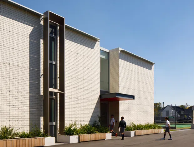 Exterior view of a contemporary, low-rise institutional or commercial building with pale cream brick walls, tall narrow glazed vertical openings framed in dark metal, and a projecting dark canopy over the entrance. Linear timber-toned planter boxes with ornamental grasses line the paved approach, while people provide scale. The building has clean rectilinear massing and a flat roofline under a clear blue sky.