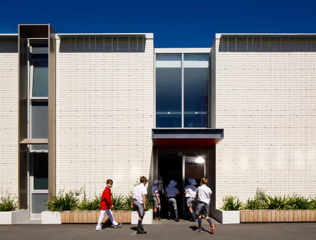 Front-facing exterior view of a modern institutional or commercial building with a symmetrical rectilinear facade. Pale textured or finely gridded wall panels flank a tall central glazed entrance, with dark horizontal canopy detailing and narrow vertical glazing at the sides. Planter beds line the base, and several people are walking near the entrance. The facade is photographed under a clear blue sky.