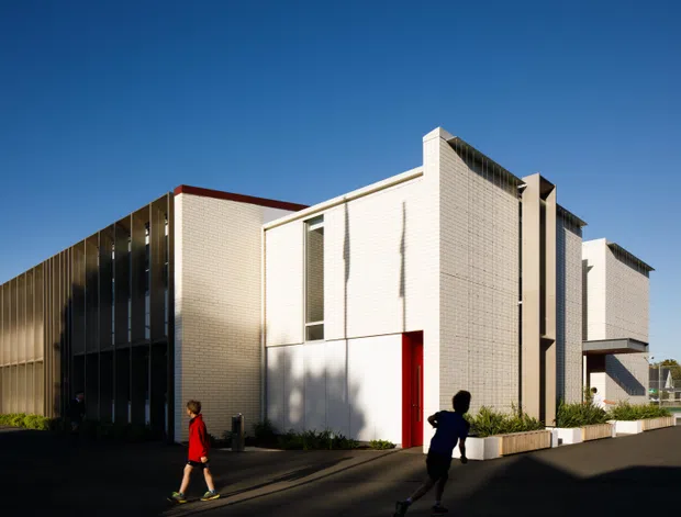 Exterior view of a contemporary multi-part institutional or commercial building with strong geometric massing. The facade combines pale brick or masonry walls, dark vertical metal screening, narrow vertical glazing, projecting white volumes, and a red entry door. The building sits along a paved pedestrian and vehicle access area under clear daylight.