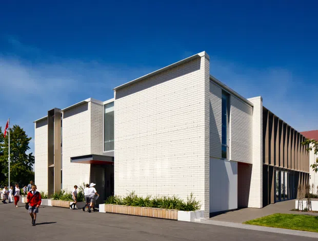 Exterior view of a contemporary institutional or commercial building with crisp geometric volumes, pale white brick walls, tall narrow glazing, and a contrasting dark vertical-screen facade. The building fronts a paved forecourt with landscaped planter beds, lawn, and people near the entrance under a clear blue sky.