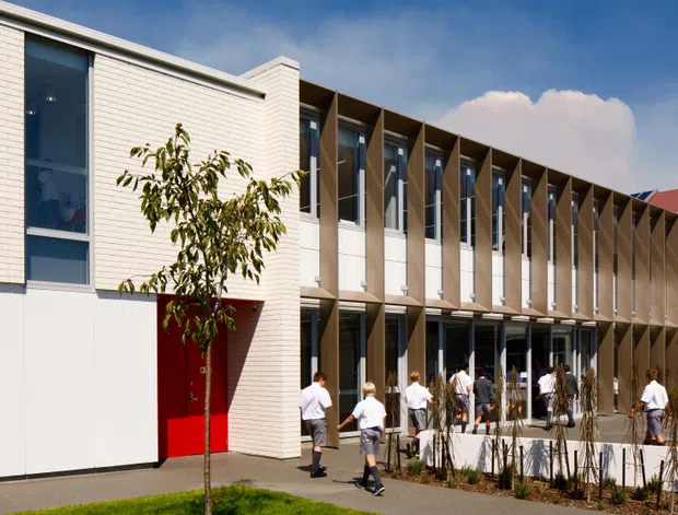 Exterior view of a contemporary multi-storey institutional or commercial building with pale brick and smooth white wall panels. A long glazed facade is articulated by repeated vertical projecting fins that provide shade and rhythm. A vivid red entrance door creates a strong accent, with students or occupants walking along a landscaped forecourt. The building has a mostly flat roofline with a small contrasting red pitched-roof section visible at the rear.