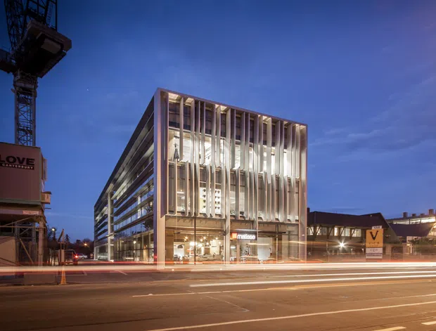Twilight exterior photograph of a contemporary multi-storey commercial building on an urban street, with a strongly illuminated glazed facade framed by dense vertical fins. Construction equipment and an adjacent building are visible, while vehicle light trails run across the foreground.