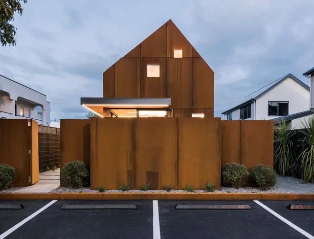 Street-facing exterior view of a compact contemporary residence with a steep gabled roof form and a dark rust-toned metal-clad facade. Small square windows punctuate the upper facade, while a projecting illuminated entry canopy sits behind a tall matching privacy fence and gate. Neat low planting and a paved pedestrian path line the frontage, with marked parking spaces in the foreground and neighbouring modern homes visible on either side.