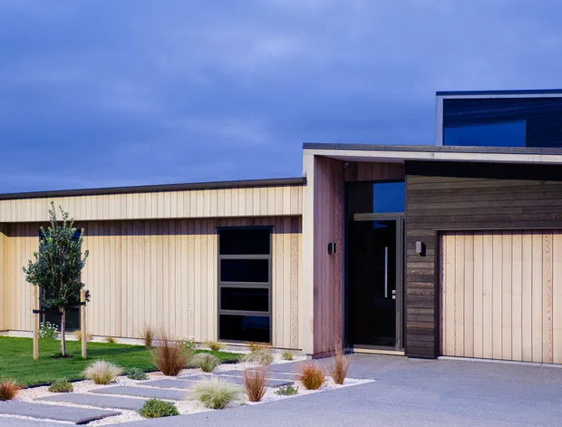 Modern single-storey residential facade photographed from the driveway, featuring a low horizontal composition, pale vertical timber cladding, darker horizontal timber cladding around the entry and garage, large dark-framed glazing, a flat roofline, and a landscaped gravel-and-paving front garden.