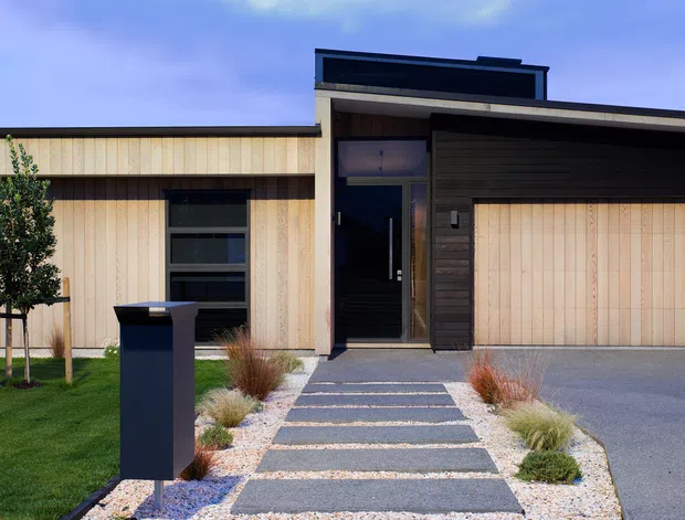 Front exterior view of a contemporary single-storey home with pale vertical timber cladding, dark contrasting entry and garage elements, a broad garage door, and a landscaped path of rectangular concrete pavers set in light gravel. The facade has clean horizontal and vertical lines, dark-framed glazing, and restrained low-maintenance planting.