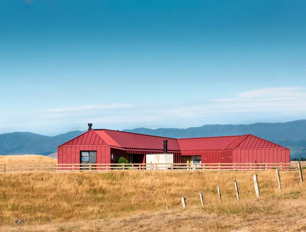 A long, single-storey rural house or farm building with bold red metal-clad walls and a matching pitched, gabled roof. The building sits behind timber fencing in a dry grassland setting, with forested mountains visible in the background beneath a clear blue sky. The central sheltered outdoor area and simple geometric form give it a contemporary agricultural or rural character.