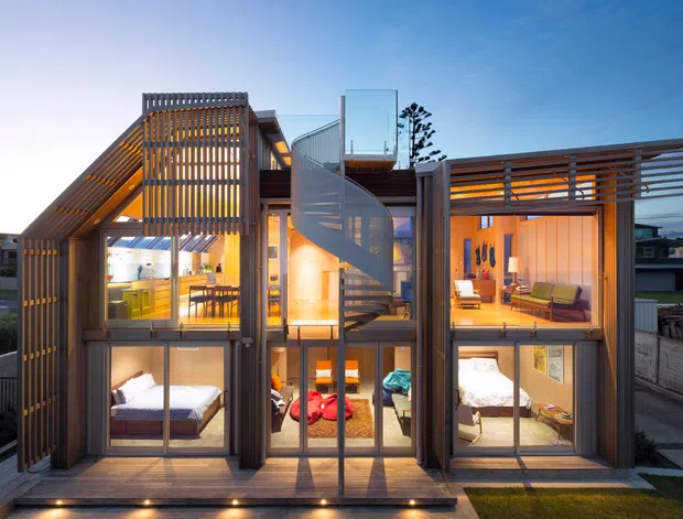 Front-facing exterior view of a contemporary multi-level timber-clad residence at dusk. The facade features extensive floor-to-ceiling glazing, vertical timber screening, horizontal timber brise-soleil, balconies, and a prominent external spiral stair. Warm interior lighting reveals a kitchen and dining area on the upper left, living spaces on the upper right, and bedrooms on the lower level. A timber deck and small landscaped foreground are visible.