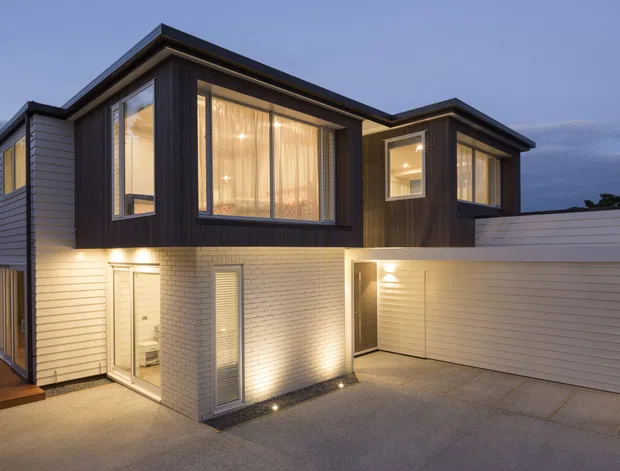 Twilight exterior view of a contemporary multi-level home with contrasting dark vertical timber-look cladding and light horizontal weatherboards, extensive glazed living areas, warm interior lighting, a projecting upper volume, and an integrated double garage. The home is approached by a paved driveway with a small timber entry deck.