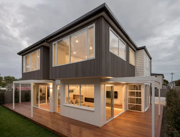 Exterior architectural photograph of a contemporary two-storey residential home at dusk. The house combines dark vertical upper-level cladding with light horizontal lower-level cladding, extensive windows, warm interior lighting, and a broad timber deck wrapping around the ground floor. A simple flat-to-low-pitched roof form and slim covered outdoor areas are visible.