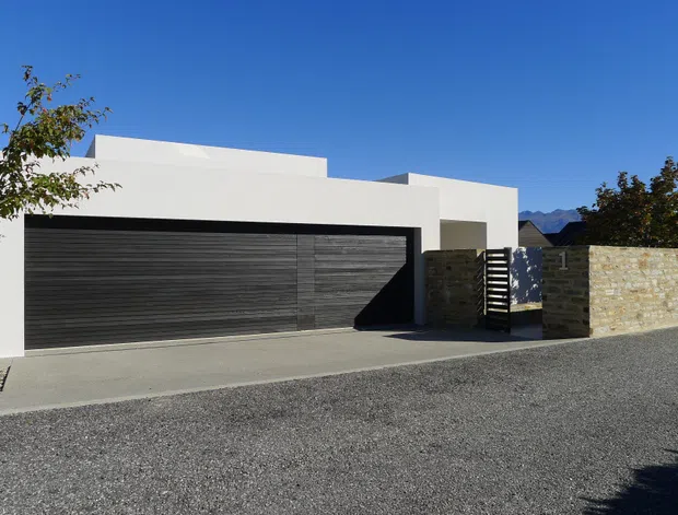 Street-facing view of a low-profile modern residence with crisp white rendered walls, a broad dark horizontal-panel garage door, a recessed entry behind a slatted screen, and a pale natural-stone boundary wall. The building has a flat roof and minimalist geometric massing, with mountains visible in the distant background.