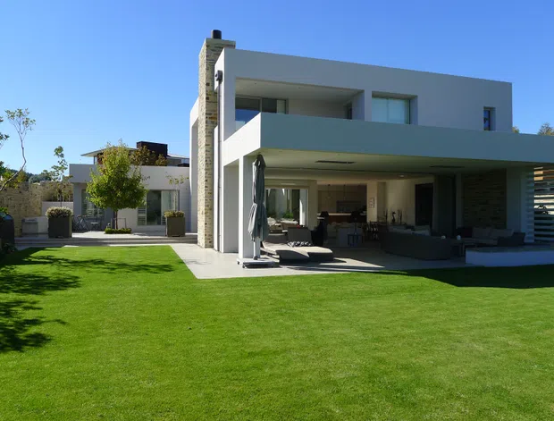 Contemporary two-storey residence with a clean white rendered facade, strong horizontal lines, recessed upper-level openings, and a broad cantilevered terrace or roof over an open outdoor living area. A tall textured masonry chimney forms a vertical counterpoint. The house opens onto a large manicured lawn, with covered outdoor seating and dining visible beneath the overhang, plus landscaped courtyard elements and privacy screening.