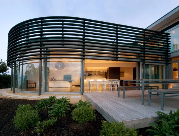 Exterior architectural photograph of a contemporary multi-level home at dusk, featuring a curved glazed living space enclosed by a dramatic horizontal black louvred screen. A timber deck with outdoor dining furniture fronts the house, while warm interior lighting reveals a living area and kitchen or bar zone behind the glazing. Landscaped planting occupies the foreground.
