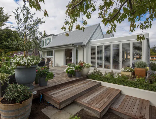 Exterior view of a light-coloured, gabled home with a steep pitched roof, enclosed porch or entry, and extensive white louvred glazing. A tiered timber deck with broad steps leads through a landscaped garden filled with potted plants and raised garden beds.