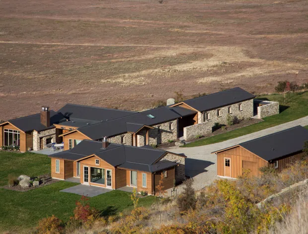 Aerial view of a sprawling single-storey rural property composed of several connected and detached buildings. The complex features dark pitched roofs, warm timber-clad wings, extensive rough stone walls, covered outdoor areas, and a separate detached garage or outbuilding. It sits within a landscaped grassy site bordered by dry native vegetation and open fields.