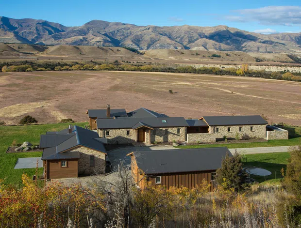 A sprawling low-rise rural residence set amid expansive cultivated fields, with rugged mountains forming the backdrop. The buildings feature dark pitched metal roofs, natural stone walls, timber-clad sections, and multiple interconnected wings. Autumnal planting surrounds the property, with a long driveway and open agricultural landscape beyond.