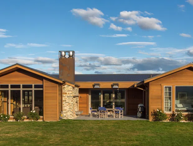 Single-storey residential home viewed from the front, composed of warm timber-clad gabled wings connected by a lower central section. The facade includes large glazed openings, stone-clad chimney elements, a covered central outdoor entertaining area, and a dark metal pitched roof. The house sits in a landscaped rural or semi-rural setting with hills in the background.