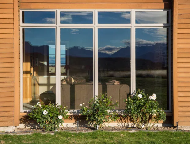 Exterior view of a single-storey building facade clad in warm horizontal timber boards, with a large multi-pane window reflecting dramatic mountain scenery. Flowering shrubs sit beneath the window, with lawn and a narrow gravel border in the foreground.