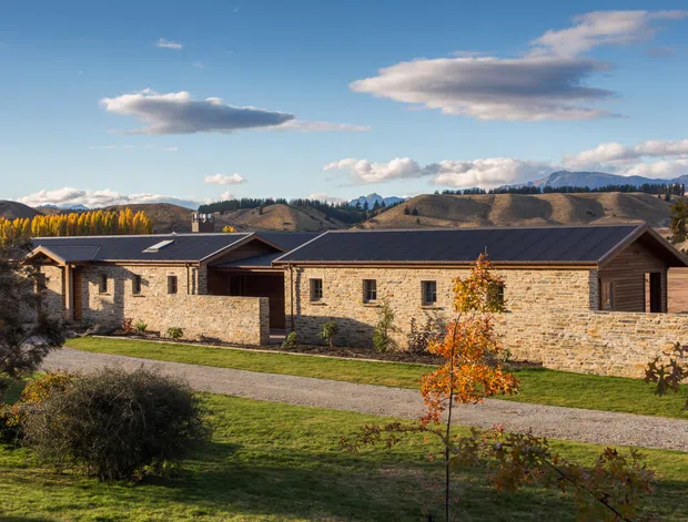 A low, single-storey stone-clad building with dark pitched roofs, set in a landscaped rural or semi-rural site. The long horizontal facade includes small rectangular windows and a recessed central entry, with rolling hills and mountains visible behind it.