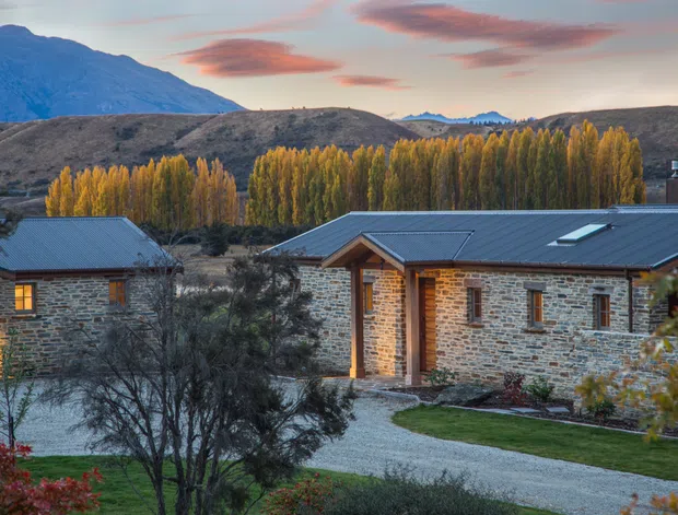 Exterior view of a low-rise stone-clad building complex set in a landscaped rural or alpine environment. The buildings have dark pitched metal roofs, timber-framed entry details, and small rectangular windows, with autumnal trees, rolling hills, and mountains in the background.
