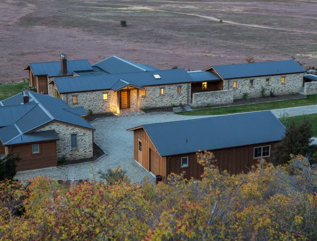Aerial view of a rural compound comprising several low-rise buildings with dark blue metal gabled roofs, natural stone walls, and warm timber-clad sections. Gravel paths and driveways connect the buildings, with autumn vegetation in the foreground and open agricultural terrain beyond.
