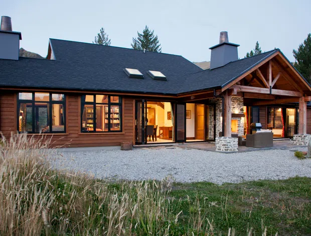 A substantial single-storey lodge-style home viewed from the front, set in a natural landscape. The building has dark timber cladding, a broad charcoal gabled roof with skylights, stone chimney bases, extensive timber-framed glazing, and a prominent covered entry or outdoor entertaining area with exposed timber trusses. Gravel landscaping and tall grasses surround the facade.