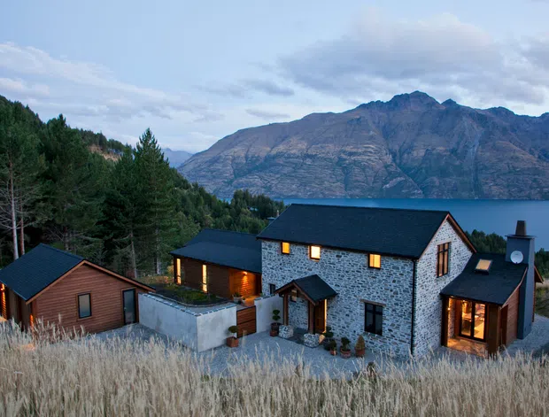A modern rustic mountain-lakeside residence viewed from above, with dark pitched roofs, stone masonry walls, timber-clad sections, warm illuminated windows, and separate connected-looking structures set among tall grasses and evergreen trees. A dramatic mountain range and lake form the background.