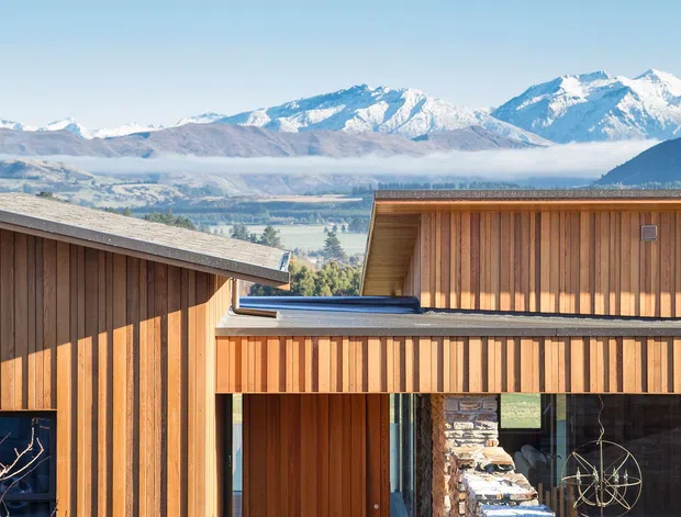 Exterior view of a contemporary mountain dwelling with warm vertical timber cladding, a low-pitched roof, large glazed openings, and a rugged natural-stone wall framing the entry courtyard. Snow-capped mountains and a broad valley form the background.