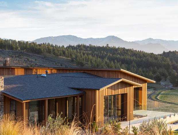 A low-profile contemporary mountain home set into a wooded, rural landscape. The building features warm vertical timber cladding, dark shingle roofing, extensive glazing, stone masonry, and broad roof overhangs. Dry grasses and a timber terrace surround the house, with forested hills and mountains visible beyond.