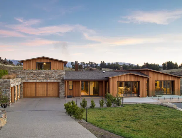 Single-storey contemporary residential home on a sloped rural or semi-rural site, composed of several low-pitched gabled timber-clad volumes. The facade includes a prominent timber garage, extensive floor-to-ceiling glazing, a covered entry, stone retaining walls, paved driveway, lawn, and landscaped planting. Warm interior lighting is visible through the windows at dusk.