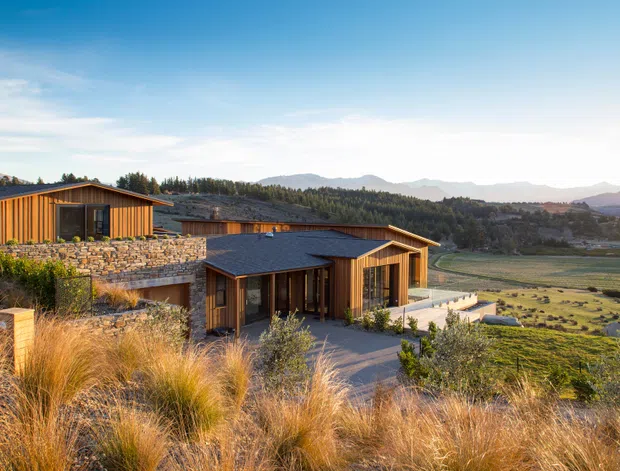 A low-profile contemporary house stepping across a sloping rural site, with warm timber-clad volumes, extensive stone retaining walls, dark pitched roofs, and broad outdoor terraces. Native grasses and rolling mountain scenery surround the home, creating a strong connection to the landscape.