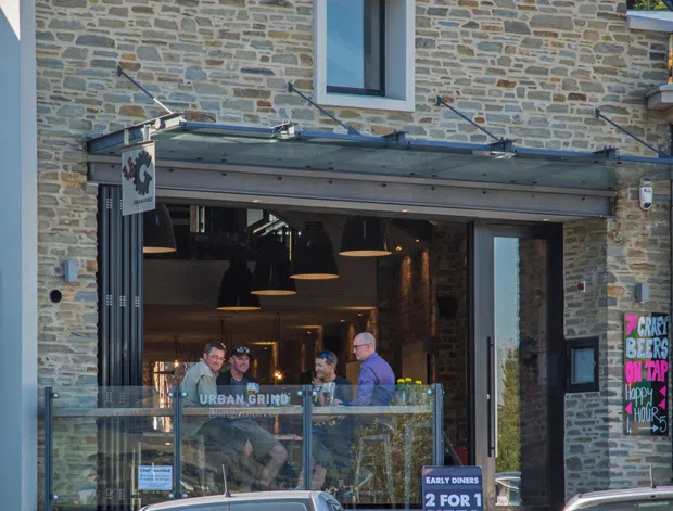 Street-facing commercial restaurant frontage in a multi-level stone-clad building. The facade features irregular grey and beige masonry, white roofline trims, a large glazed storefront entrance, a projecting glass canopy, exterior signage, and an open dining area visible inside. Cars and people are present in the street foreground and interior.