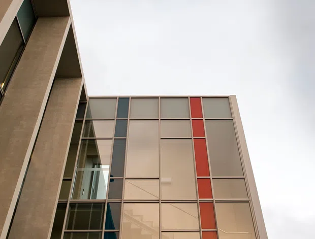 Upward-looking exterior architectural photograph of a contemporary multi-storey building. The facade combines tall concrete side walls with a large rectilinear curtain-wall glazing system, opaque translucent panels, and narrow vertical red accent panels. Interior staircases and circulation spaces are visible behind the glazing under an overcast sky.